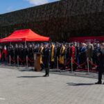 Military ceremony with uniformed officers standing in formation behind a red carpet and podium under a red canopy.