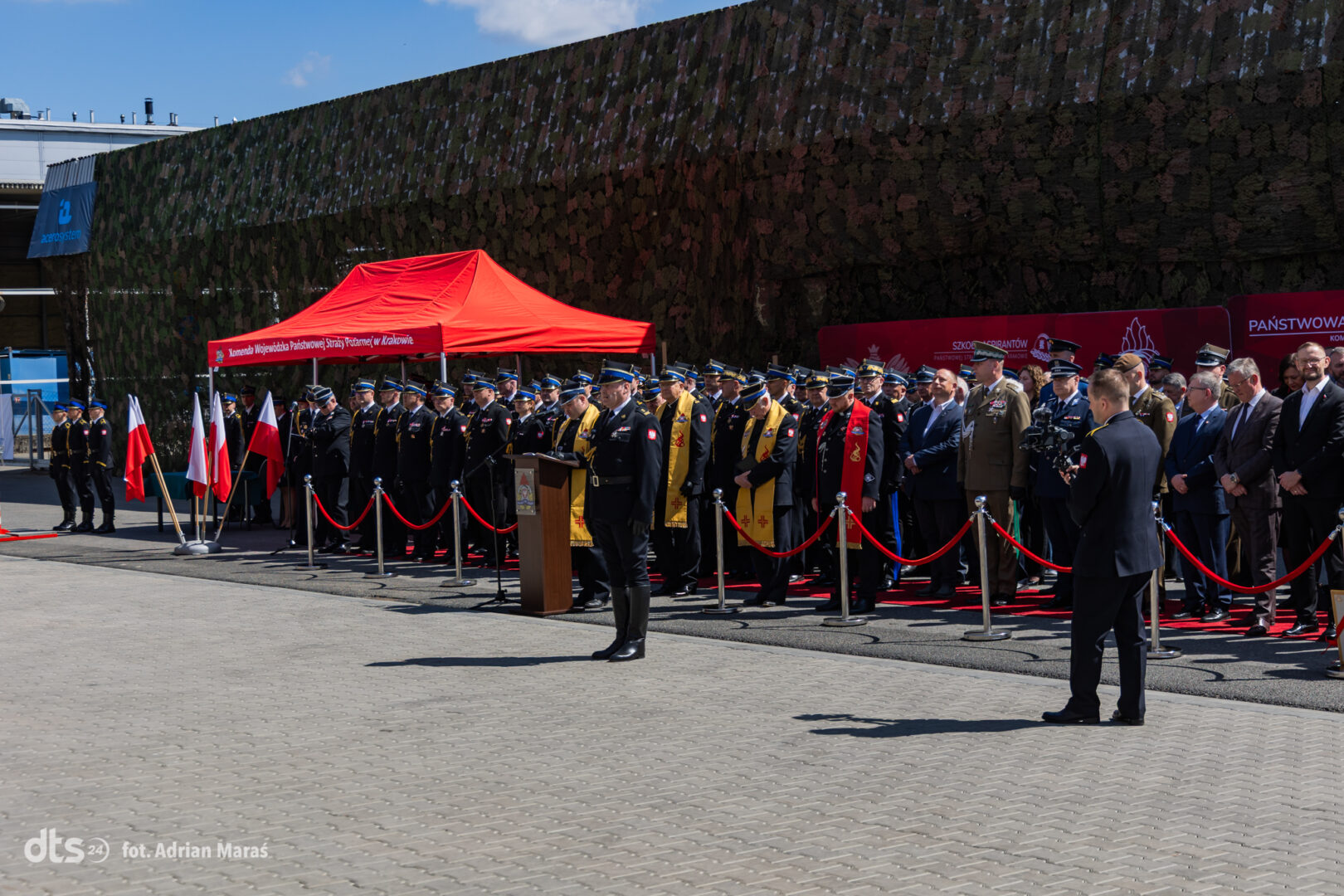 Military ceremony with uniformed officers standing in formation behind a red carpet and podium under a red canopy.