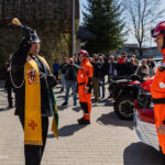 Priest blessing rescue workers with holy water as crowd watches, orange-clad responders and emergency vehicles in the background.