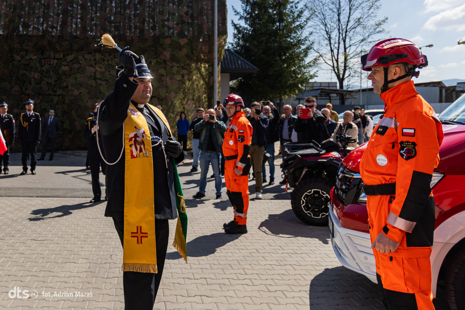Priest blessing rescue workers with holy water as crowd watches, orange-clad responders and emergency vehicles in the background.