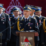 Central military officer in dress uniform speaks into a microphone at a podium during a ceremony, with medals and a rope epaulette visible.