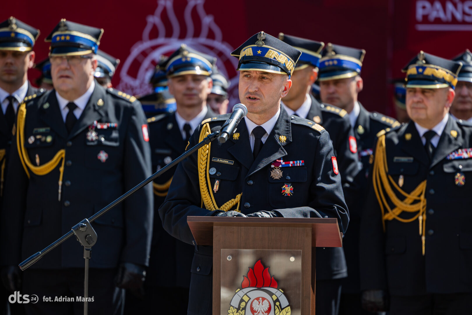 Central military officer in dress uniform speaks into a microphone at a podium during a ceremony, with medals and a rope epaulette visible.