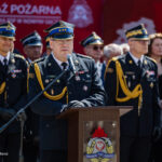 Senior firefighter officer delivering a speech at a podium during a formal ceremony, with other uniformed officers behind him on a sunny day.
