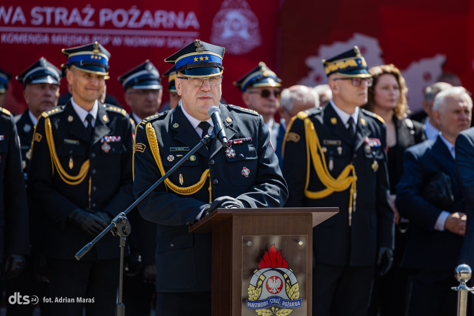 Senior firefighter officer delivering a speech at a podium during a formal ceremony, with other uniformed officers behind him on a sunny day.