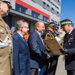 Military officers in dress uniforms shake hands with a man in a suit as a framed award is presented outdoors at a formal ceremony.