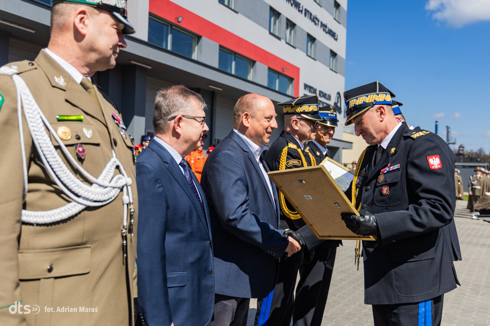 Military officers in dress uniforms shake hands with a man in a suit as a framed award is presented outdoors at a formal ceremony.