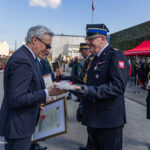 Two men in formal attire exchange a framed certificate at a military ceremony, with a uniformed officer presenting a wrapped item as soldiers stand in formation nearby.