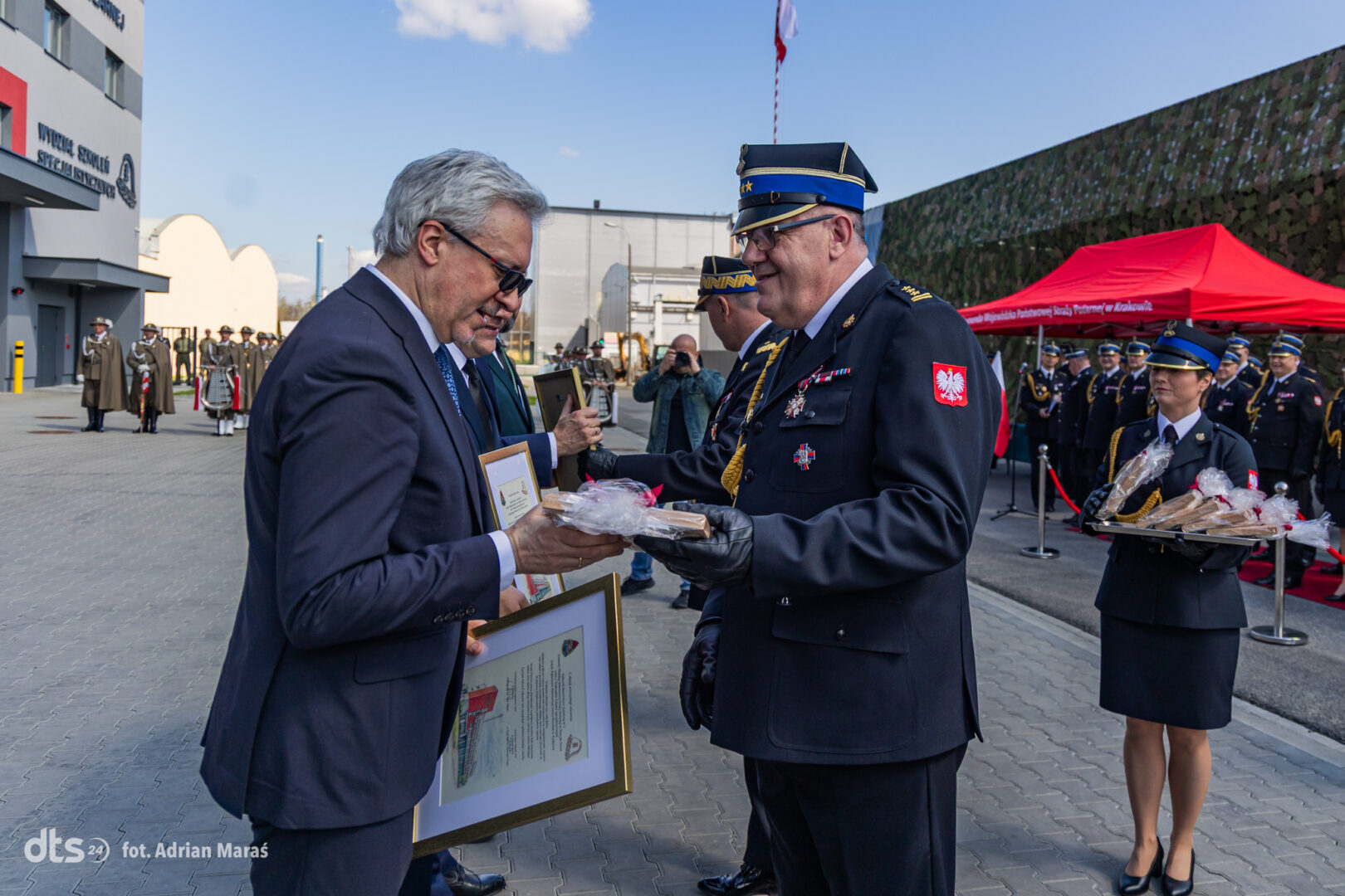 Two men in formal attire exchange a framed certificate at a military ceremony, with a uniformed officer presenting a wrapped item as soldiers stand in formation nearby.
