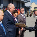 Uniformed officer hands a plaque to formally dressed civilians during an outdoor ceremony, with soldiers lined up in the background.
