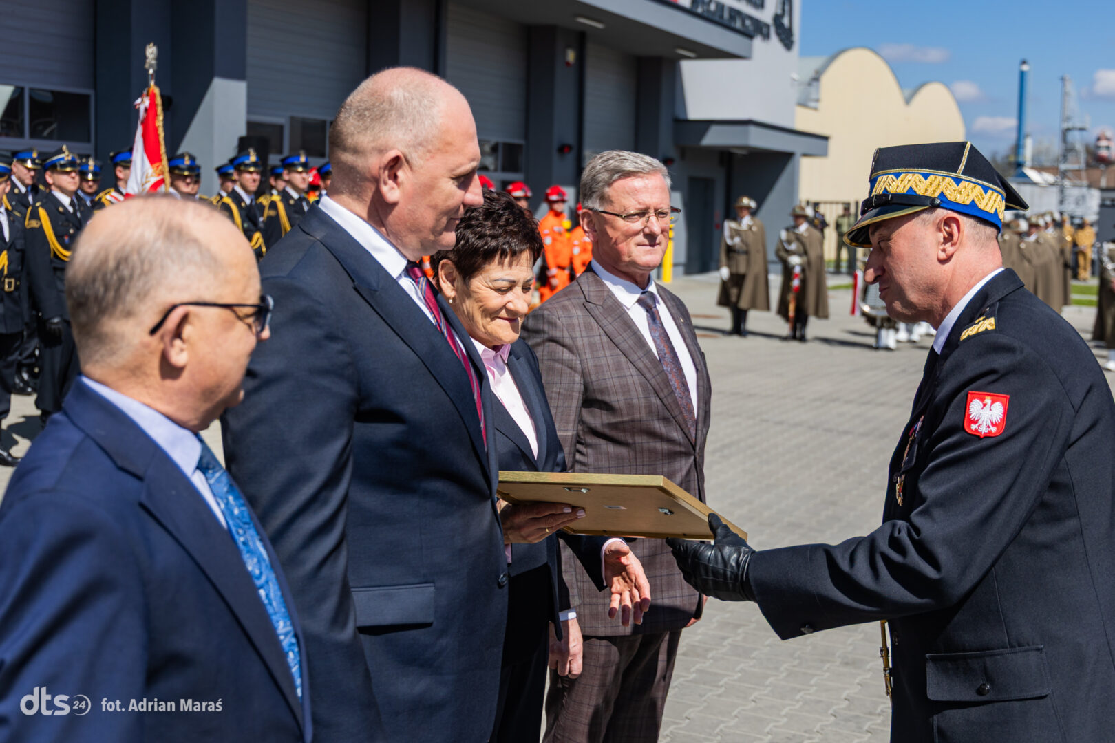 Uniformed officer hands a plaque to formally dressed civilians during an outdoor ceremony, with soldiers lined up in the background.