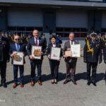 Five award recipients stand side by side outdoors, each holding a framed certificate and small gift, with uniformed personnel in the background.