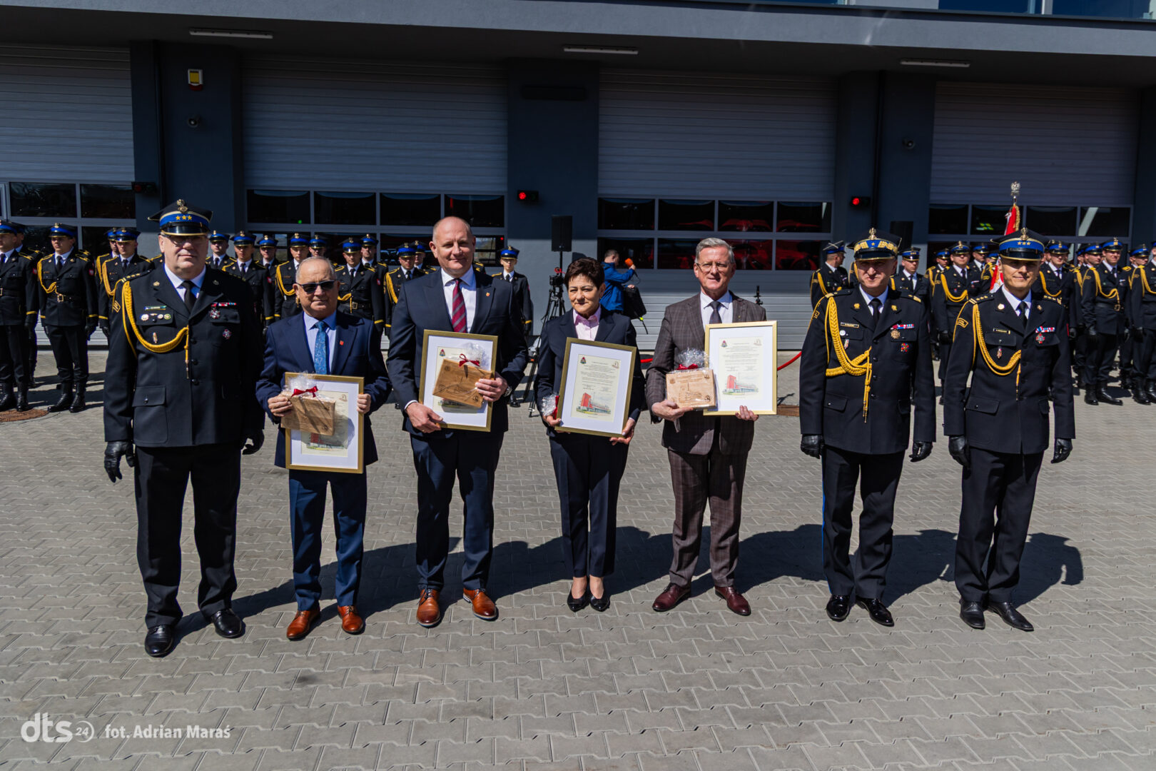 Five award recipients stand side by side outdoors, each holding a framed certificate and small gift, with uniformed personnel in the background.