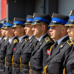 Row of soldiers in black ceremonial uniforms with blue peaked caps standing in formation during a parade, gold cords visible on jackets.