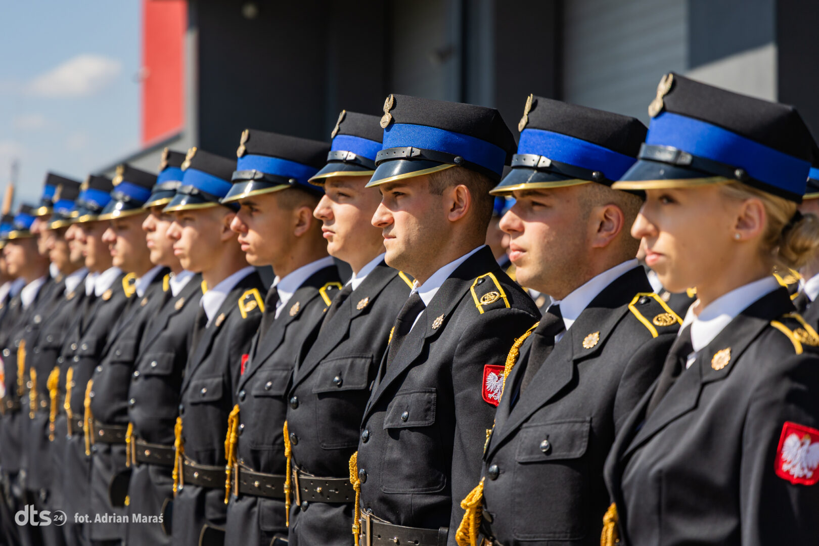 Row of soldiers in black ceremonial uniforms with blue peaked caps standing in formation during a parade, gold cords visible on jackets.