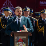 Man in a teal suit speaks at a lectern with microphone; military officers stand behind at a formal ceremony.