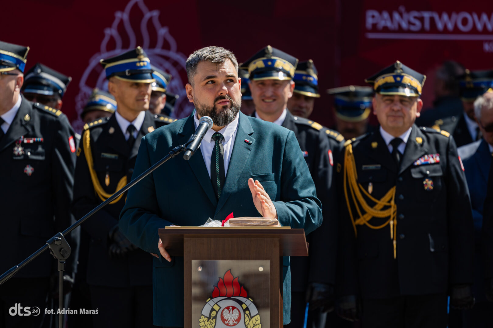 Man in a teal suit speaks at a lectern with microphone; military officers stand behind at a formal ceremony.