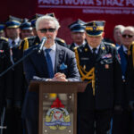 Man in a dark suit and sunglasses speaks at a wooden podium with a microphone, flanked by formally dressed firefighters in black uniforms and gold braided epaulettes.