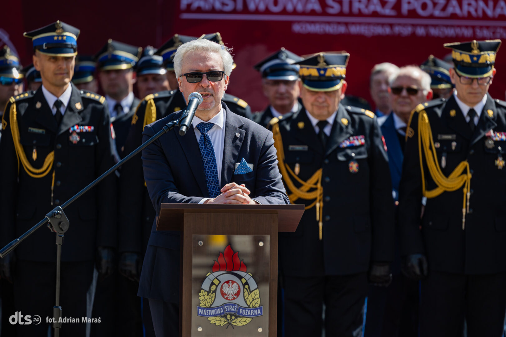 Man in a dark suit and sunglasses speaks at a wooden podium with a microphone, flanked by formally dressed firefighters in black uniforms and gold braided epaulettes.