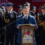Man in a blue coat speaks at a wooden podium with a microphone, fire service officers stand behind him at a ceremony