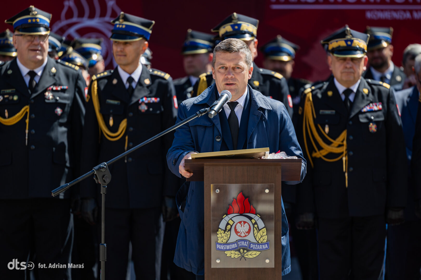 Man in a blue coat speaks at a wooden podium with a microphone, fire service officers stand behind him at a ceremony