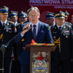 Man in a blue suit speaks into a microphone at a podium during a formal ceremony, with uniformed firefighters behind him.