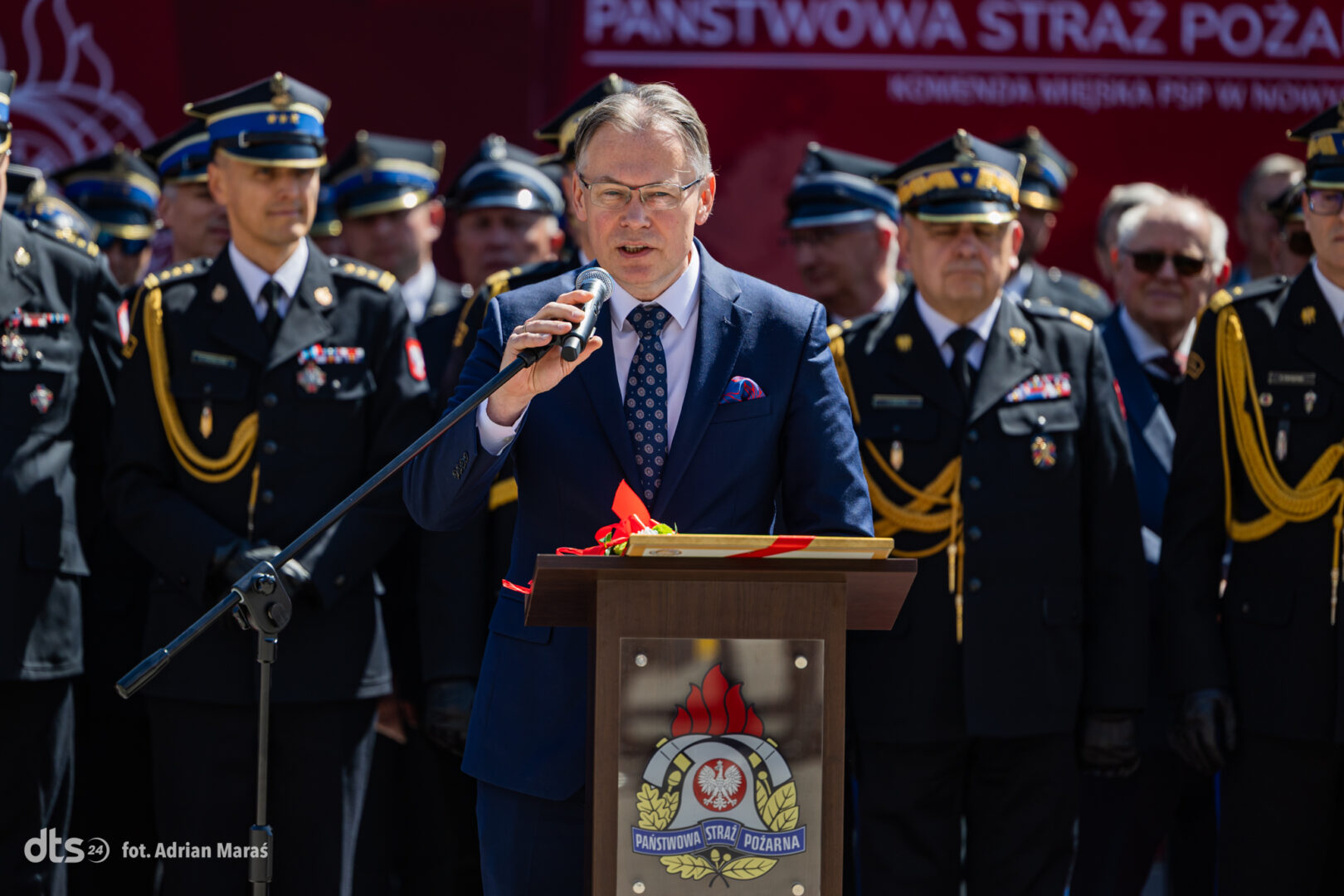 Man in a blue suit speaks into a microphone at a podium during a formal ceremony, with uniformed firefighters behind him.