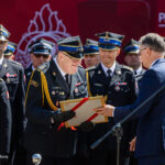 Senior officer in ceremonial dress receives a certificate from a man in a blue suit during an award ceremony, with fellow officers in uniform in the background.