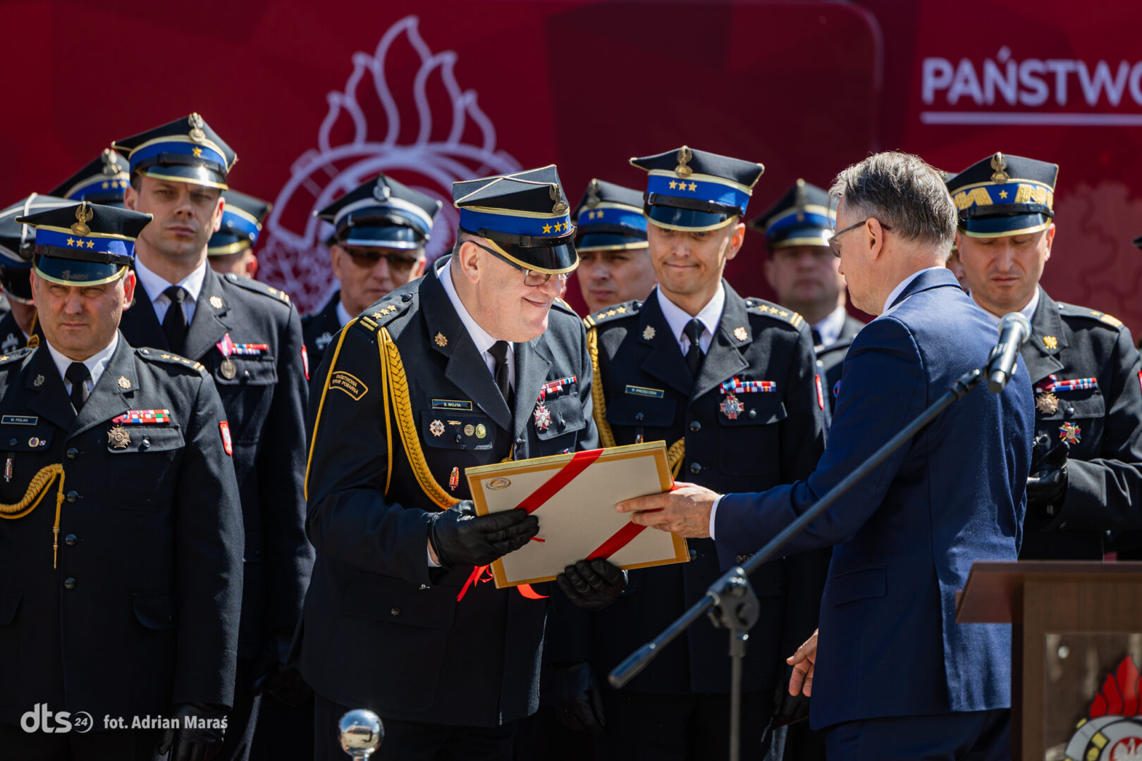 Senior officer in ceremonial dress receives a certificate from a man in a blue suit during an award ceremony, with fellow officers in uniform in the background.