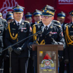 Senior officer in ceremonial uniform speaks at a podium, flanked by uniformed colleagues during a formal ceremony.