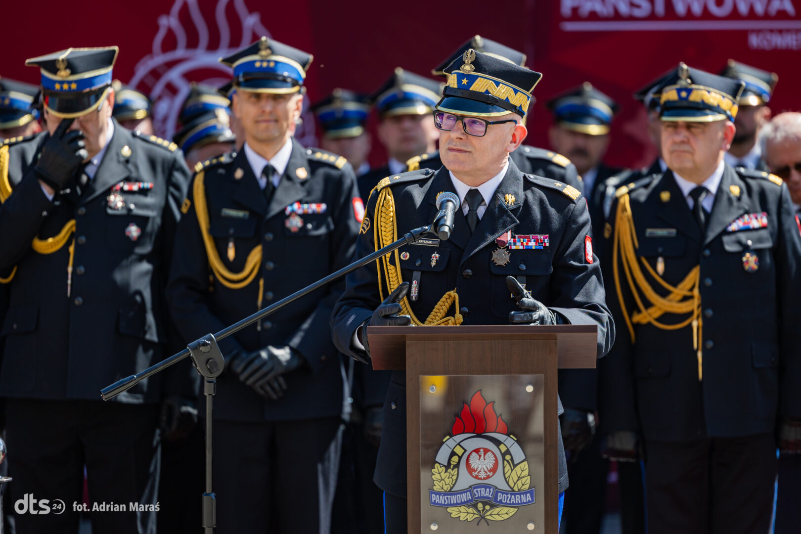 Senior officer in ceremonial uniform speaks at a podium, flanked by uniformed colleagues during a formal ceremony.