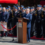 Speaker at a wooden podium addressing a formal military ceremony, with uniformed officers lined up behind and red carpet in view.