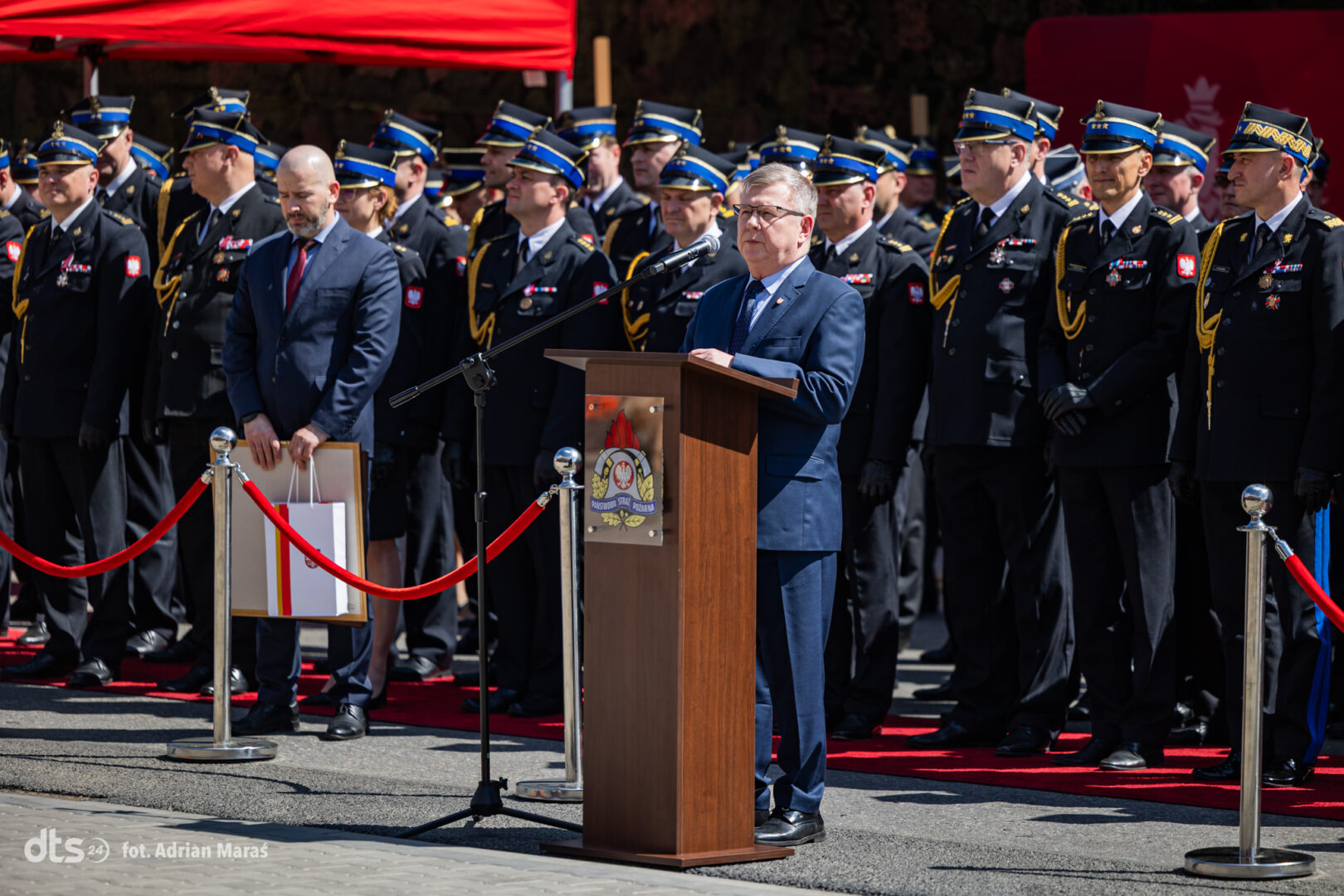 Speaker at a wooden podium addressing a formal military ceremony, with uniformed officers lined up behind and red carpet in view.