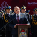 Speaker in a dark suit at a podium with a fire service emblem, flanked by uniformed officers at a formal ceremony.