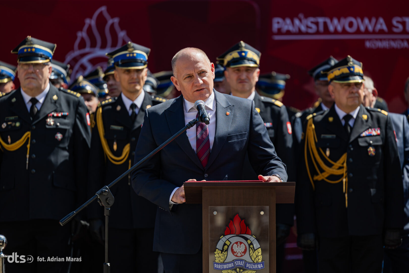 Speaker in a dark suit at a podium with a fire service emblem, flanked by uniformed officers at a formal ceremony.