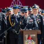 Military officer at a podium delivering a speech, surrounded by uniformed colleagues during a ceremony.
