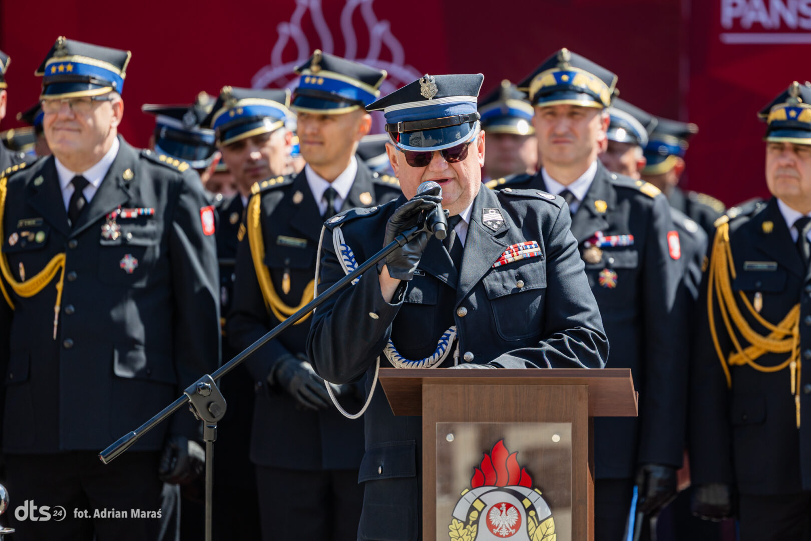 Military officer at a podium delivering a speech, surrounded by uniformed colleagues during a ceremony.