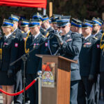 Senior officer in dress uniform giving a speech at a podium during a formal military ceremony, with a row of uniformed personnel behind him.