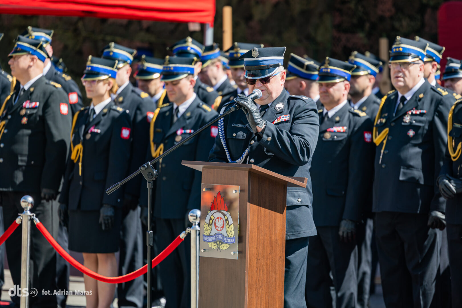 Senior officer in dress uniform giving a speech at a podium during a formal military ceremony, with a row of uniformed personnel behind him.