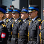 Line of uniformed soldiers standing at attention in black dress uniforms with blue peaked caps and gold trim, Poland patches visible on sleeves
