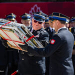 Senior officer in a peaked cap hands a bundle of framed certificates tied with red ribbons to a colleague at a formal ceremony, with other uniformed officers in the background.