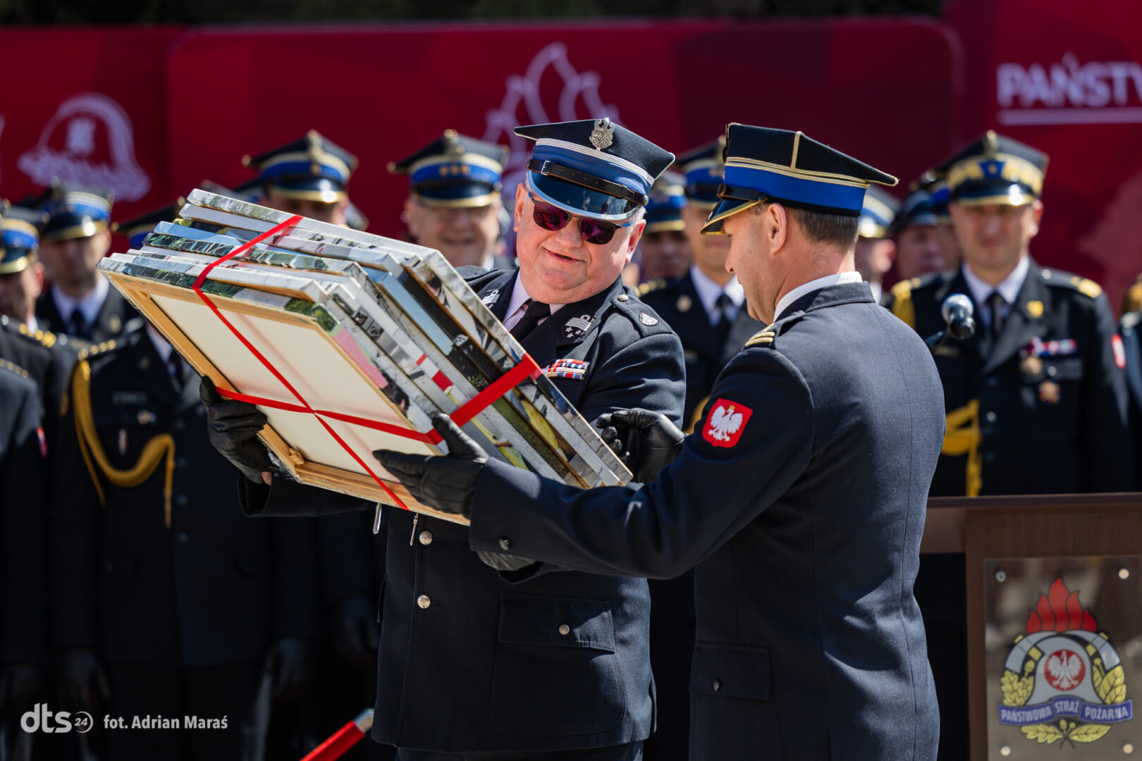 Senior officer in a peaked cap hands a bundle of framed certificates tied with red ribbons to a colleague at a formal ceremony, with other uniformed officers in the background.