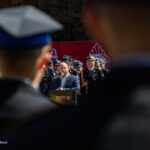 Man in a suit delivers a speech at a podium during a military ceremony, with uniformed officers standing behind him.