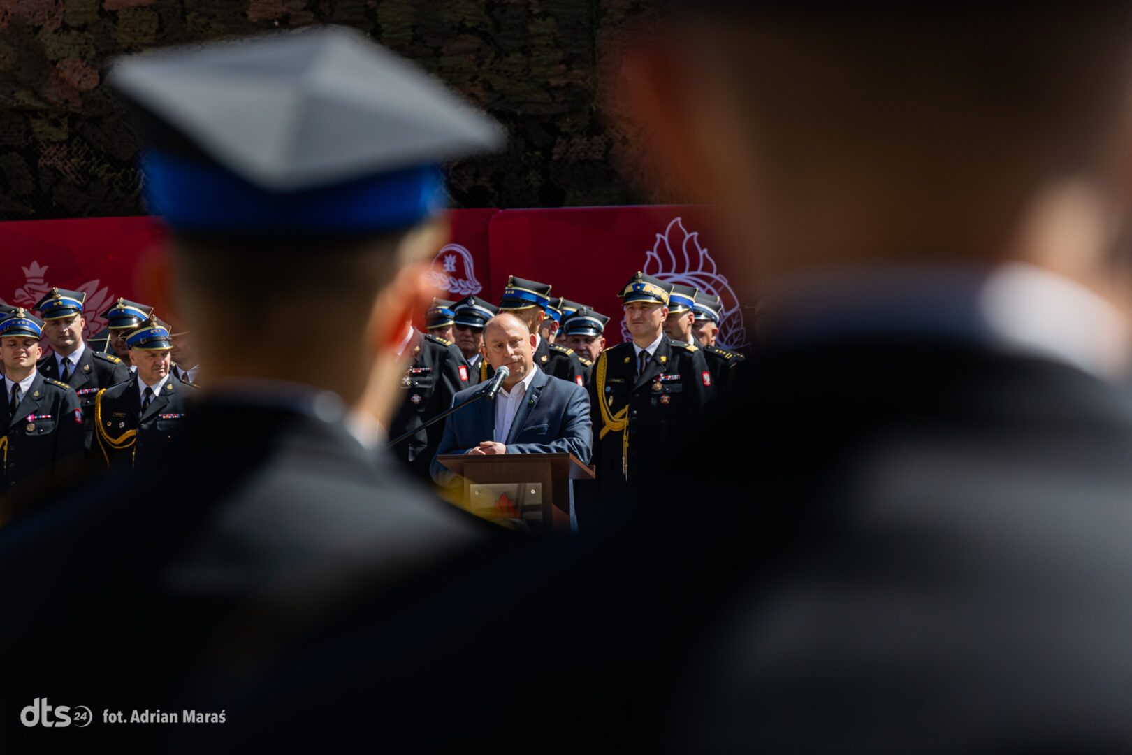 Man in a suit delivers a speech at a podium during a military ceremony, with uniformed officers standing behind him.