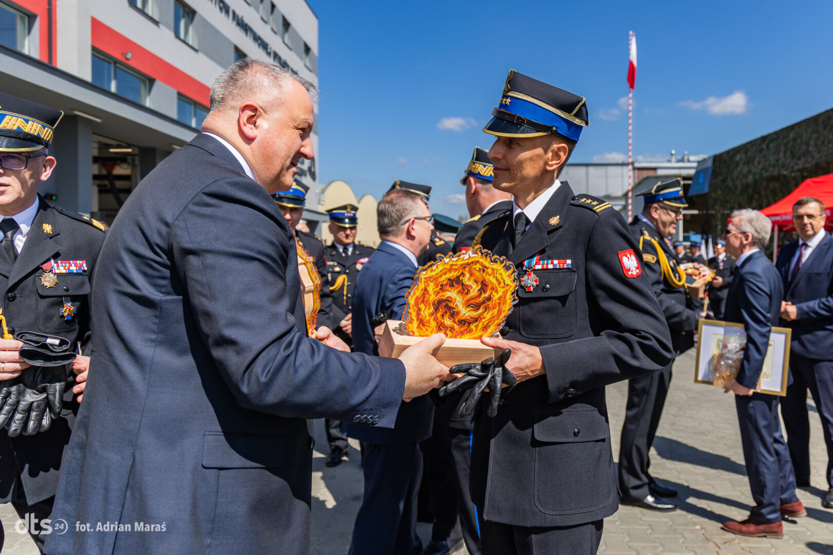 Two uniformed officers exchange a ceremonial flaming emblem during an outdoor military awards event, with other soldiers and spectators in the background.