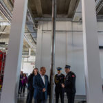 Group of people in business and military attire stand around a tall polished metal column in a modern industrial space outside a construction or showroom area.
