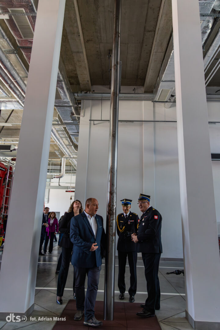 Group of people in business and military attire stand around a tall polished metal column in a modern industrial space outside a construction or showroom area.