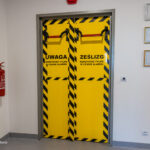 Yellow double emergency door with black diagonal hazard stripes and red horizontal bars; Polish warning to use only during an alarm.