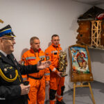 Speaker in a formal uniform addresses two rescue workers in orange gear, who hold a statue beside a damaged brick display and a religious painting on an easel.