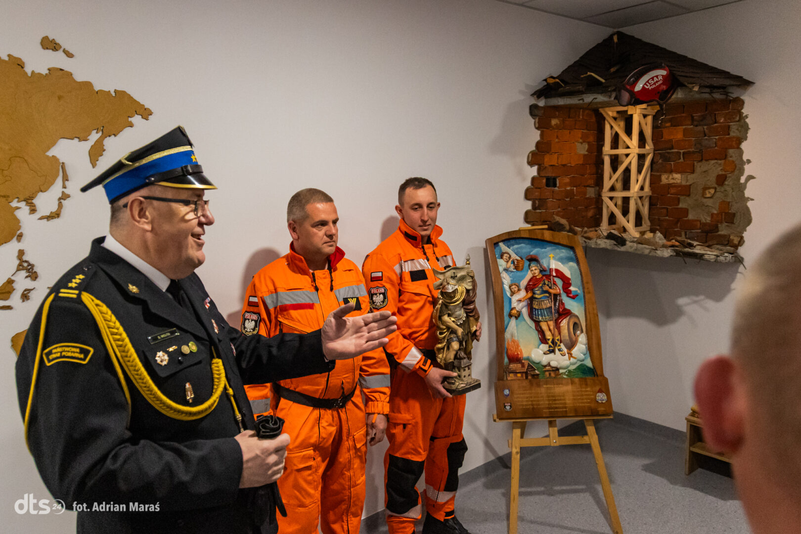 Speaker in a formal uniform addresses two rescue workers in orange gear, who hold a statue beside a damaged brick display and a religious painting on an easel.
