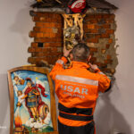 USAR Poland firefighter in an orange uniform reattaching a statue atop rubble, as a photographer records the scene on the right.